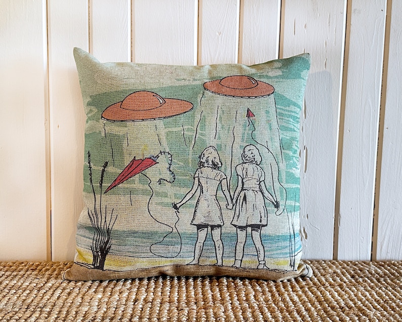 May include: A green and brown throw pillow with a vintage illustration of two girls standing in front of a beach scene with flying saucers and a red kite. The pillow has a light brown border.