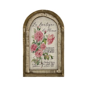 May include: A rustic wooden framed sign with a white background and a floral design. The sign reads "La Boutique de Fleurs" and features pink roses with green leaves. The sign has a distressed, vintage look.