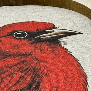 May include: Close-up of a red bird with a black eye and a white beak. The bird is depicted on a canvas with a brown wooden frame.