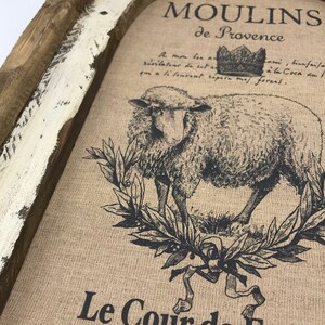 Op de afbeelding: Een rustiek houten frame met een witte verweerde afwerking omringt een jute canvas print. De print toont een zwart-witte illustratie van een schaap met een kroon erboven. De tekst "MOULINS de Provence" en "Le Cour de Ferm" is op het canvas gedrukt.