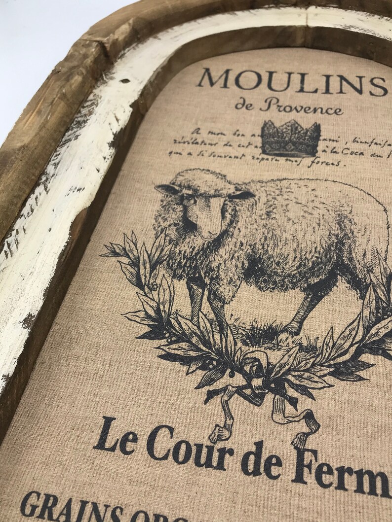 Op de afbeelding: Een rustiek houten frame met een witte verweerde afwerking omringt een jute canvas print. De print toont een zwart-witte illustratie van een schaap met een kroon erboven. De tekst "MOULINS de Provence" en "Le Cour de Ferm" is op het canvas gedrukt.
