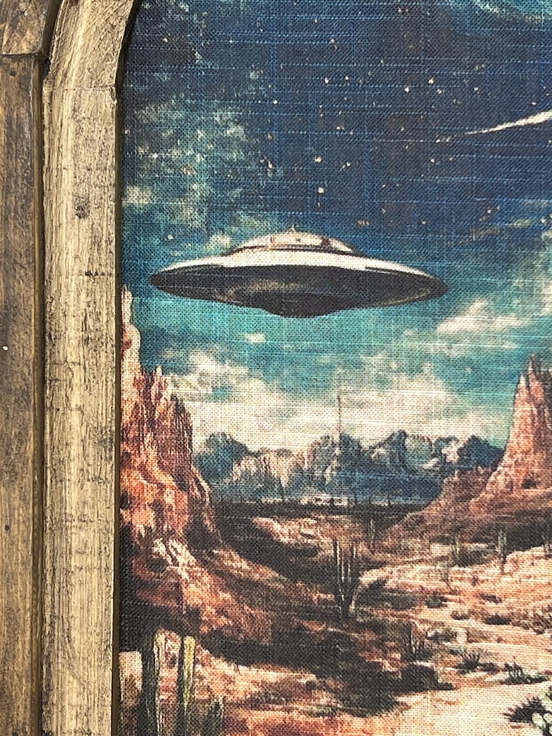 May include: A framed painting depicting a desert landscape with a flying saucer hovering over a canyon. The sky is a deep blue with stars and a comet. The desert is a warm brown with cacti and mountains in the distance.