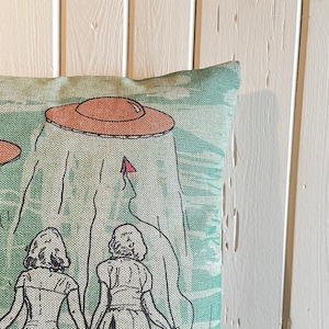 May include: A green and white throw pillow with a design of two women standing under two flying saucers. The saucers are orange and brown.