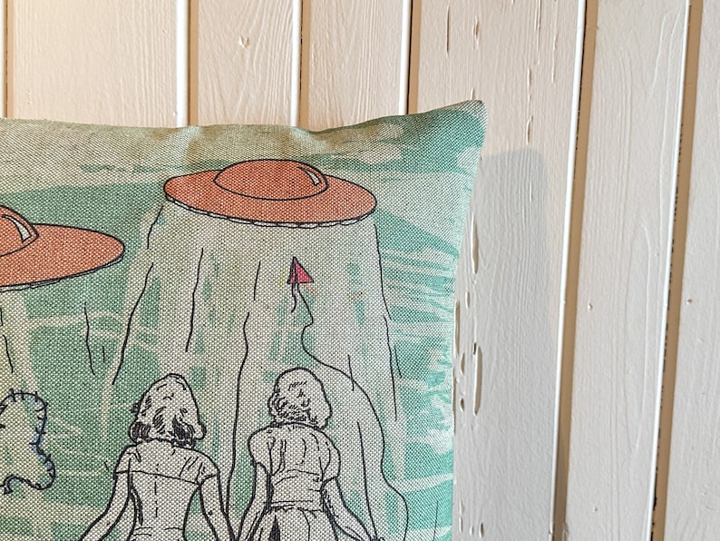 May include: A green and white throw pillow with a design of two women standing under two flying saucers. The saucers are orange and brown.