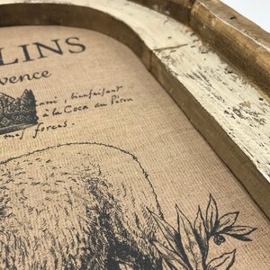 Op de afbeelding: Een ingelijste print met een rustieke houten lijst. De print toont een zwart-witte illustratie van een beer met een kroon op zijn kop en de tekst "LINS" en "vence" boven de beer. De tekst onder de beer luidt "ami, bienfaisant a la Coca du feron forces."