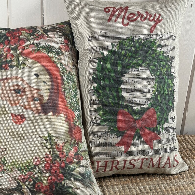May include: Two decorative throw pillows with a Christmas theme. One pillow features a vintage illustration of Santa Claus with holly and berries. The other pillow has a green wreath with a red bow on a background of sheet music with the words "Merry Christmas".