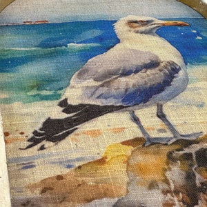 Op de afbeelding: Een aquarel schilderij van een meeuw die op een rots zit met de oceaan op de achtergrond. De meeuw is wit met zwarte vleugels en een gele snavel. De oceaan is blauw met witte golven. De rots is bruin en heeft een textuur.