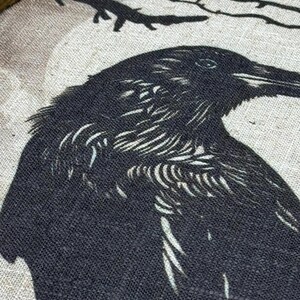 May include: A black crow with a detailed, textured design is printed on a beige canvas background. The crow is facing the right side of the image and has a sharp beak. The background is a light beige with a subtle texture.