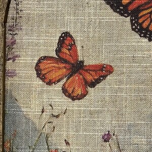 May include: A framed canvas print featuring orange and black butterflies with detailed wing patterns. The artwork includes a field of wildflowers in shades of pink, red, and purple, set against a light beige textured background.