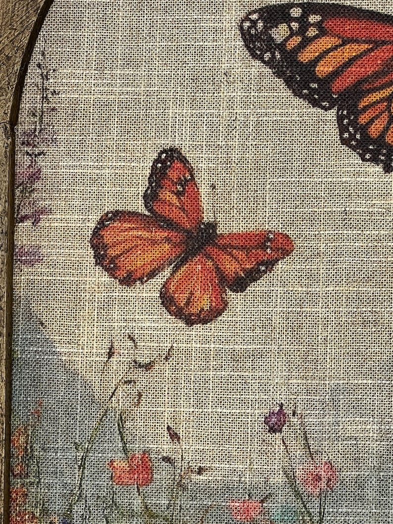 May include: A framed canvas print featuring orange and black butterflies with detailed wing patterns. The artwork includes a field of wildflowers in shades of pink, red, and purple, set against a light beige textured background.