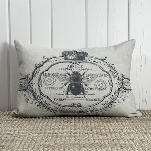 May include: A beige linen pillow with a black and white vintage design featuring a bee, a crown, and French text that reads "Abeille Constant Crepeaux Le Monant Let Ses Reves Lettres Et Jo La Montai Aux Professeur Par Grenoble En Volume In- Paris France".