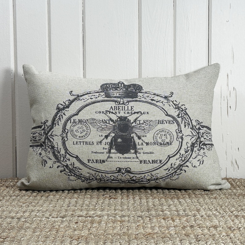 May include: A beige linen pillow with a black and white vintage design featuring a bee, a crown, and French text that reads "Abeille Constant Crepeaux Le Monant Let Ses Reves Lettres Et Jo La Montai Aux Professeur Par Grenoble En Volume In- Paris France".