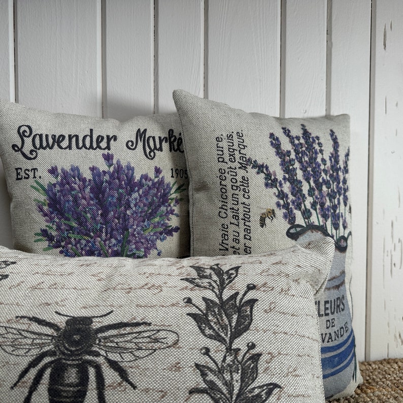 May include: Three decorative throw pillows with floral and bee designs. The pillows are made of a light beige fabric with a subtle texture. The first pillow features a bouquet of purple lavender with the text "Lavender Market Est. 1905". The second pillow features a bouquet of purple lavender with French text. The third pillow features a black and white illustration of a bee and a sprig of leaves.