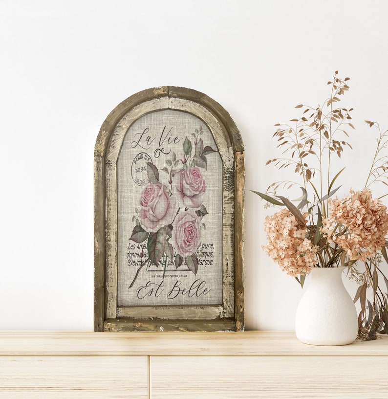 Pu&ograve; includere: Una stampa d'arte incorniciata con un'estetica vintage. La stampa presenta un motivo floreale con rose rosa e il testo "La Vie Est Belle" in un carattere decorativo. La cornice &egrave; in legno con una finitura bianca invecchiata.