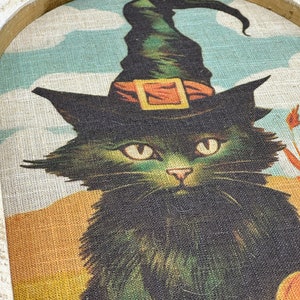 May include: A black cat wearing a green witch's hat with an orange buckle. The cat is looking directly at the viewer. The background is a light blue and yellow.