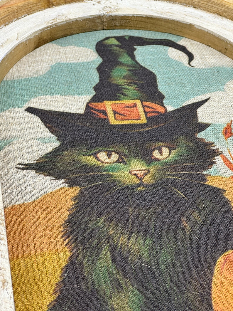 May include: A black cat wearing a green witch's hat with an orange buckle. The cat is looking directly at the viewer. The background is a light blue and yellow.