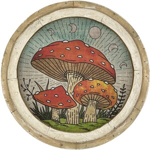 May include: A round wooden wall decor with a white frame and a vintage-style illustration of three mushrooms with red and orange caps. The mushrooms are surrounded by green leaves and a blue background with white crescent moons.