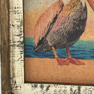 May include: A framed print of a pelican standing on a beach with a sunset in the background. The frame is made of wood and has a white distressed finish.