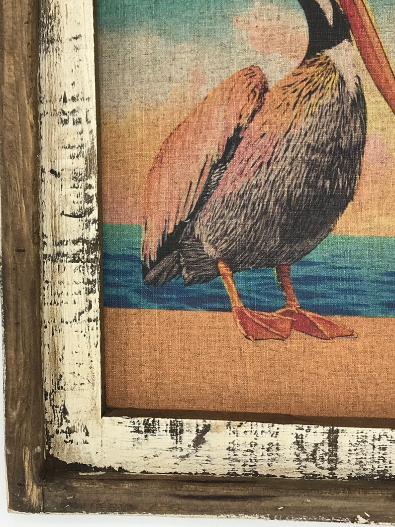 May include: A framed print of a pelican standing on a beach with a sunset in the background. The frame is made of wood and has a white distressed finish.