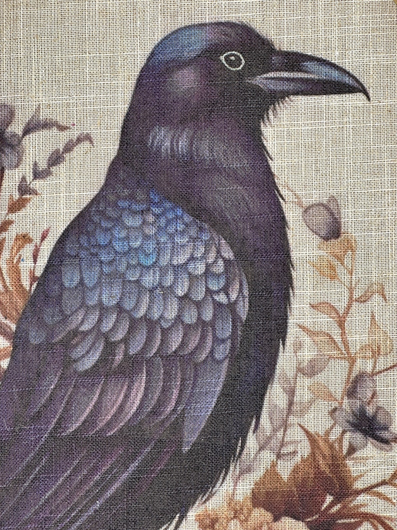 May include: A black raven with iridescent blue feathers is perched on a branch with brown and white flowers. The raven's eye is visible and the background is a beige linen fabric.