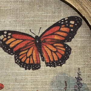 May include: A framed artwork featuring a monarch butterfly with orange, black, and yellow wings. The butterfly is centered on a textured, beige background, with floral accents in the lower right corner. The frame is a natural wood color.