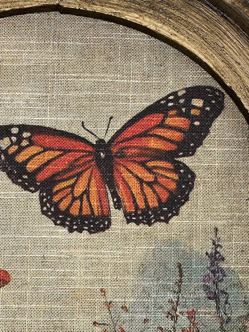 May include: A framed artwork featuring a monarch butterfly with orange, black, and yellow wings. The butterfly is centered on a textured, beige background, with floral accents in the lower right corner. The frame is a natural wood color.