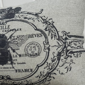 May include: A beige pillow with a black and white design featuring a bee, a stamp, and the text "ET SES REVES LA MONTAGNE FRANCE".