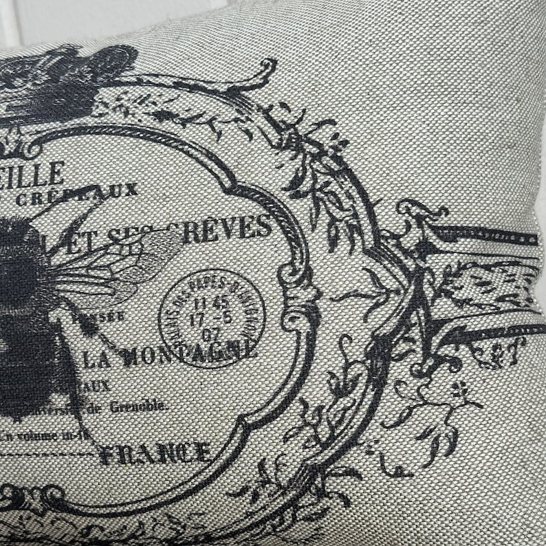 May include: A beige pillow with a black and white design featuring a bee, a stamp, and the text "ET SES REVES LA MONTAGNE FRANCE".