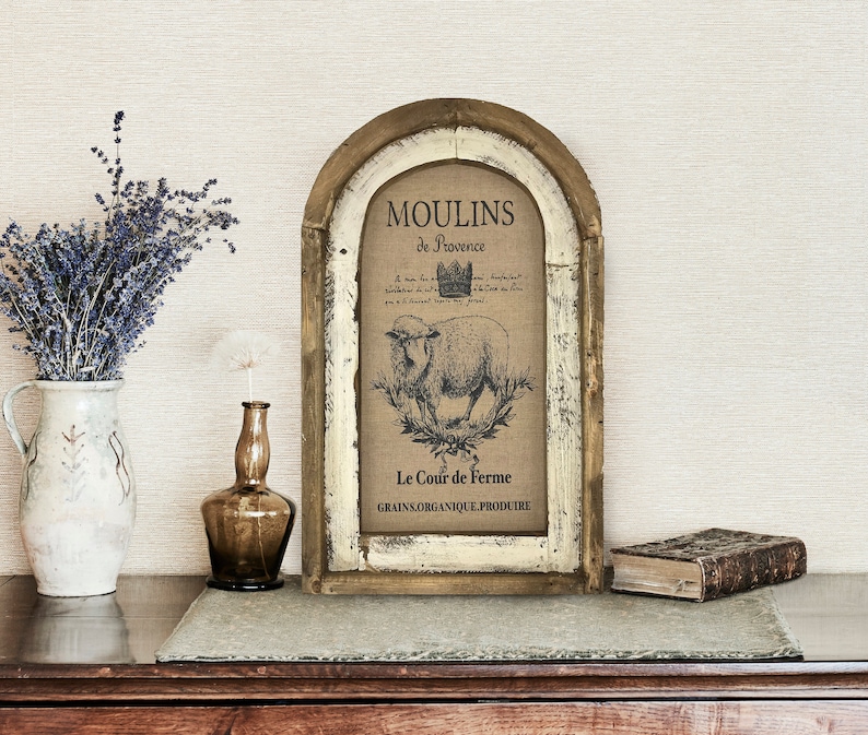 Op de afbeelding: Een rustiek houten frame met een verweerde witte afwerking, met een vintage print van een schaap met de tekst "Moulins de Provence Le Cour de Ferme GRAINS ORGANIQUE PRODUIRE".