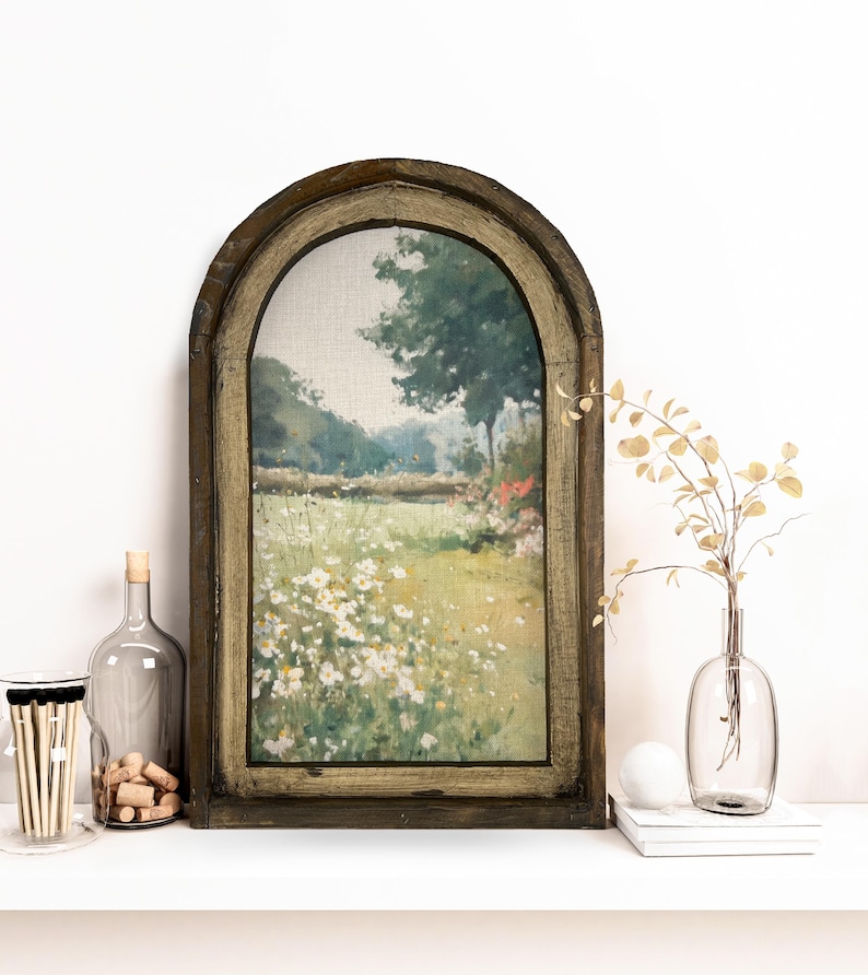 May include: An arched wooden framed landscape painting featuring a field of white flowers. A clear glass bottle with a cork stopper, a glass jar with matches, and a clear vase with dried stems are on a white shelf.