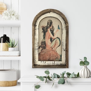 May include: A framed print of a mermaid with long flowing hair, sitting on a rock and holding a blue butterfly. The frame is made of wood with a distressed white finish.