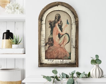 May include: A framed print of a mermaid with long flowing hair, sitting on a rock and holding a blue butterfly. The frame is made of wood with a distressed white finish.