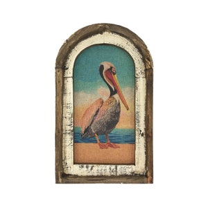 May include: A framed print of a pelican standing on a beach with a blue ocean in the background. The frame is made of wood with a whitewashed finish.