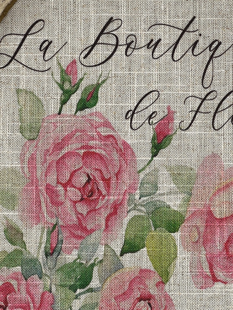 May include: A burlap fabric background with a floral design featuring pink roses and green leaves. The text "La Boutique de Fleurs" is written in black cursive script.
