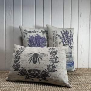 May include: Three decorative throw pillows with a floral and bee design. The pillows are made of a light beige linen fabric. The first pillow has the text "Lavender Market Est. 1905" printed on it. The second pillow has a black and white illustration of a bee and a wreath of leaves. The third pillow has a floral design with the text "Fleurs de Lavande" printed on it.