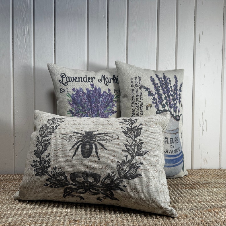 May include: Three decorative throw pillows with a floral and bee design. The pillows are made of a light beige linen fabric. The first pillow has the text "Lavender Market Est. 1905" printed on it. The second pillow has a black and white illustration of a bee and a wreath of leaves. The third pillow has a floral design with the text "Fleurs de Lavande" printed on it.