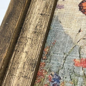 May include: Close-up of a framed floral print. The frame is made of distressed, dark brown wood. The print features a colorful garden scene with red, blue, and purple flowers, and a butterfly. The artwork is on a textured, light-colored fabric.
