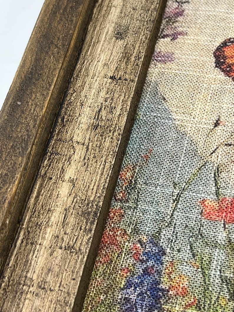 May include: Close-up of a framed floral print. The frame is made of distressed, dark brown wood. The print features a colorful garden scene with red, blue, and purple flowers, and a butterfly. The artwork is on a textured, light-colored fabric.