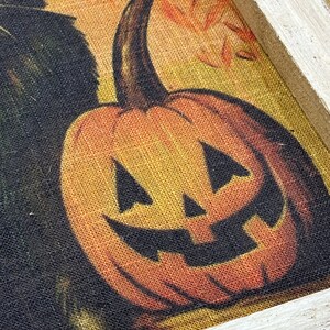 May include: A framed burlap print of a jack-o'-lantern with a toothy grin. The pumpkin is orange with black eyes and a mouth. The background is a yellow and orange burlap with a black cat peeking out from the left side.