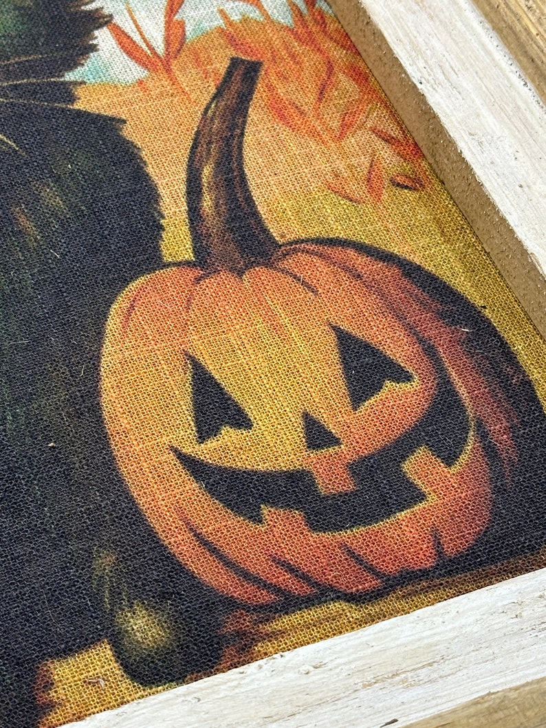 May include: A framed burlap print of a jack-o'-lantern with a toothy grin. The pumpkin is orange with black eyes and a mouth. The background is a yellow and orange burlap with a black cat peeking out from the left side.