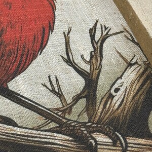 May include: A close-up of a red bird perched on a branch. The bird is depicted in a stylized, almost cartoon-like manner, with a thick, rounded body and a short, stubby beak. The branch is brown and gnarled, with a few smaller branches extending from it. The background is a light beige, suggesting a canvas or fabric surface.