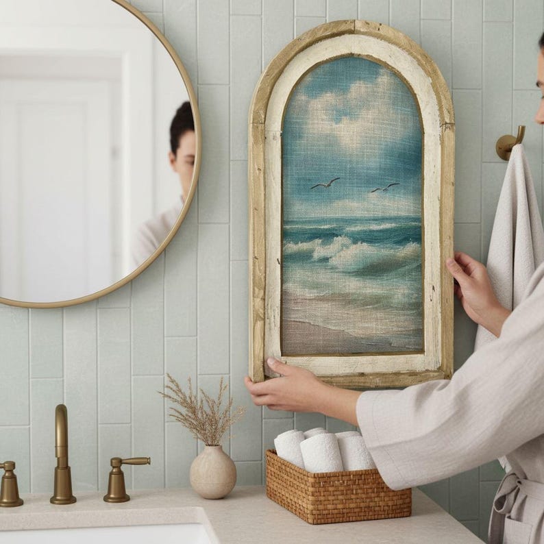 A person is holding a framed painting of a seascape in a bathroom, with a round mirror and a basket of towels nearby.
