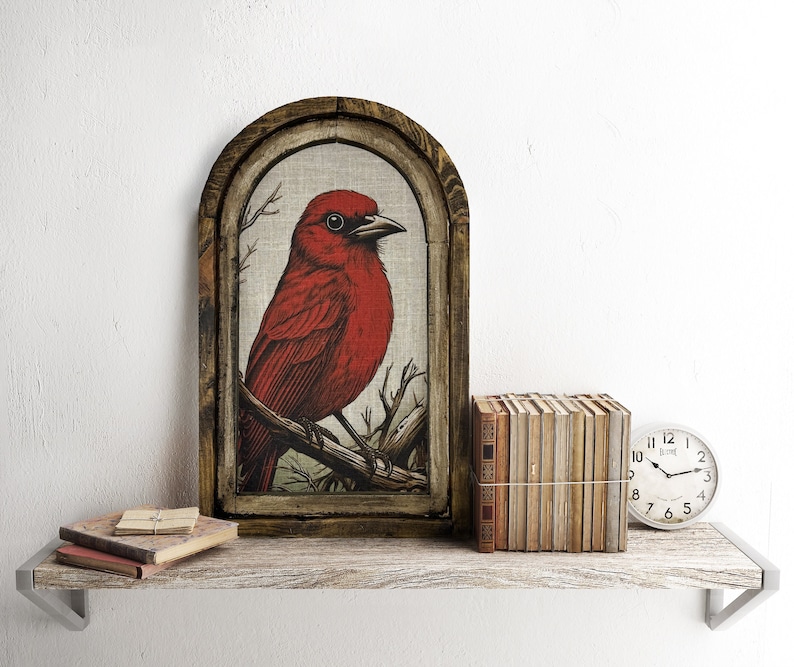 May include: A framed print of a red bird with a brown wooden frame. The print is on a linen-like material and the bird is perched on a branch. The frame is on a wooden shelf with a stack of books and a clock.