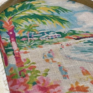 K&ouml;nnte beinhalten: Ein gerahmtes Kunstwerk mit einer tropischen Szene mit Palmen, einem Strand und einem Haus in der Ferne. Das Kunstwerk befindet sich auf einem Jute-Hintergrund mit Blumenmustern.