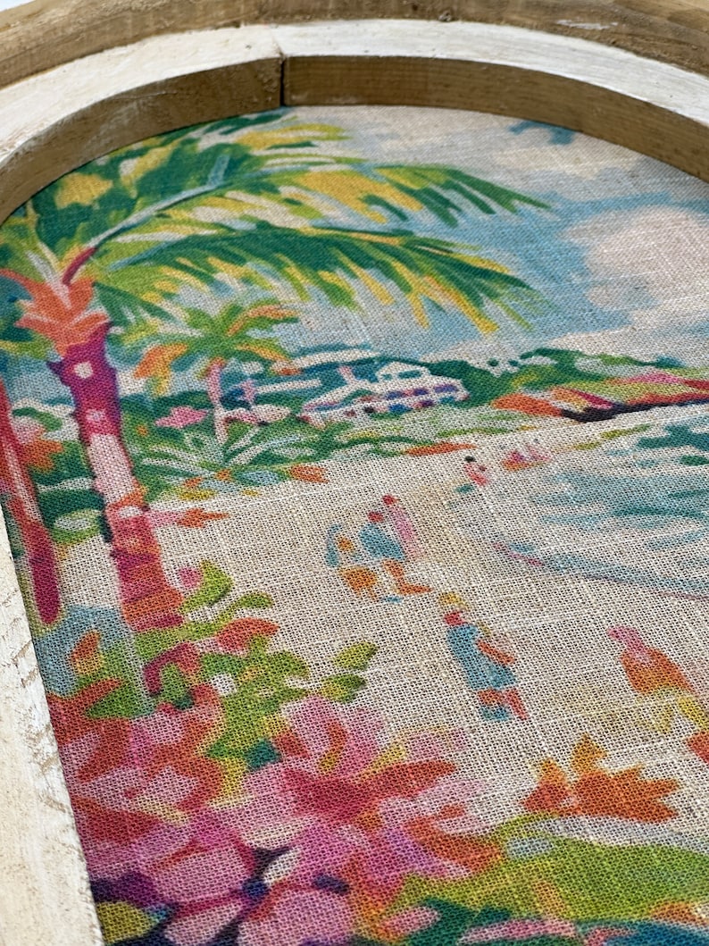 K&ouml;nnte beinhalten: Ein gerahmtes Kunstwerk mit einer tropischen Szene mit Palmen, einem Strand und einem Haus in der Ferne. Das Kunstwerk befindet sich auf einem Jute-Hintergrund mit Blumenmustern.