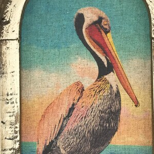 May include: A brown and white pelican with a long orange beak stands on a beach with blue water in the background. The pelican is in a framed picture with a white frame.
