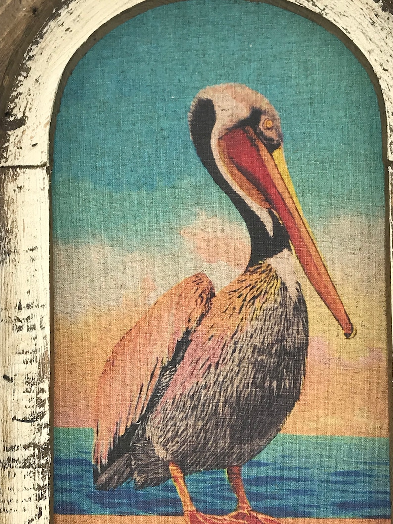 May include: A brown and white pelican with a long orange beak stands on a beach with blue water in the background. The pelican is in a framed picture with a white frame.