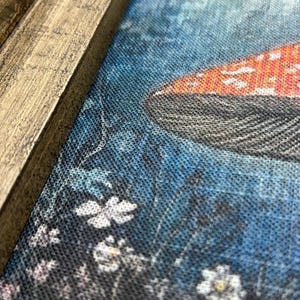May include: Close-up of a canvas print featuring a mushroom with a red cap and white spots. The background is a blue and black floral design. The print is framed in a rustic, light brown wooden frame.