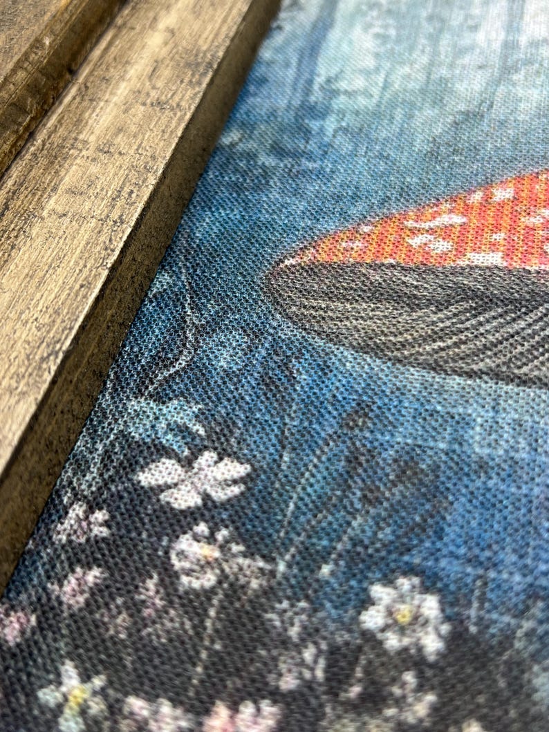 May include: Close-up of a canvas print featuring a mushroom with a red cap and white spots. The background is a blue and black floral design. The print is framed in a rustic, light brown wooden frame.