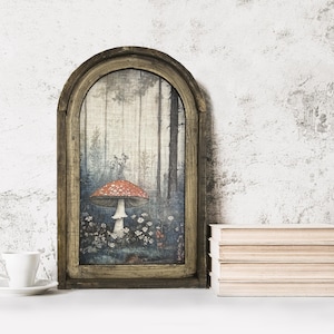 May include: A framed art print featuring a forest scene with a large red and white mushroom. The artwork is set in a rustic, arched wooden frame. A white teacup and saucer are in the foreground, with a stack of books to the right.